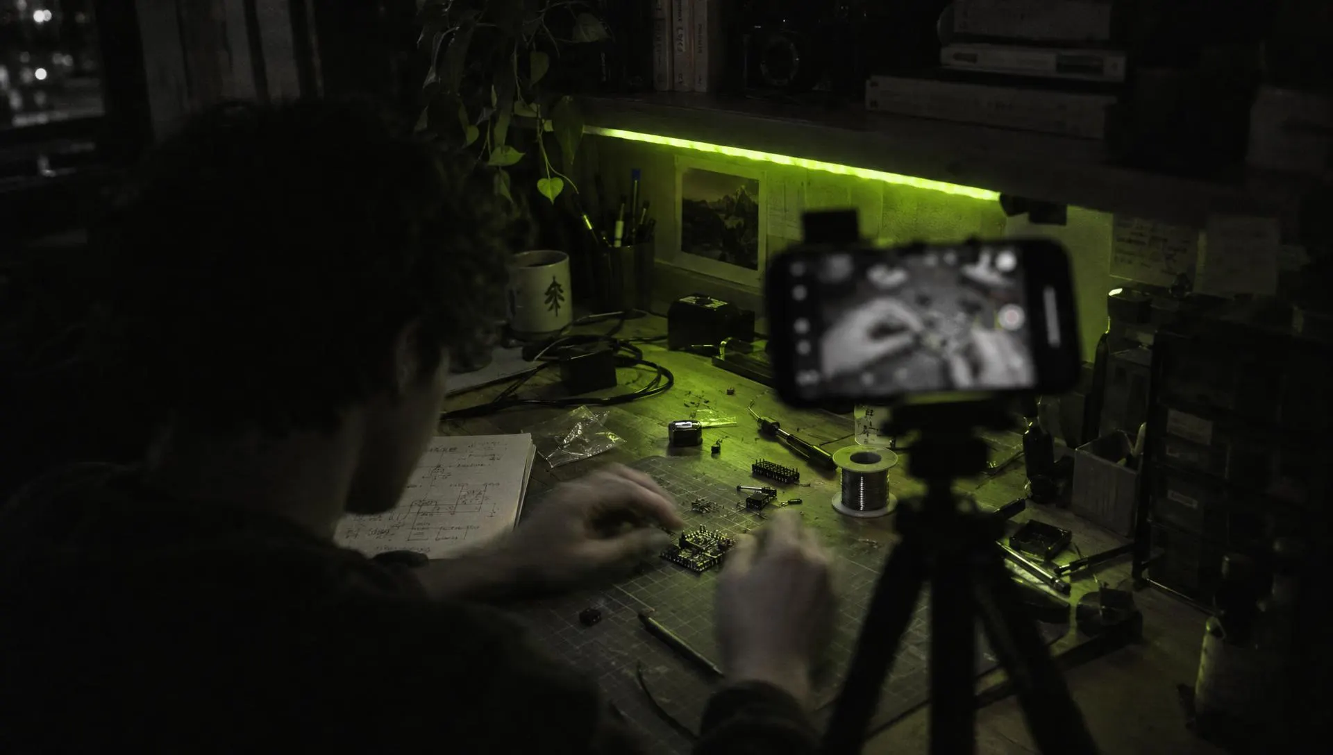 A maker filming their own desk at night, a phone on a small tripod, a green LED strip on the shelf