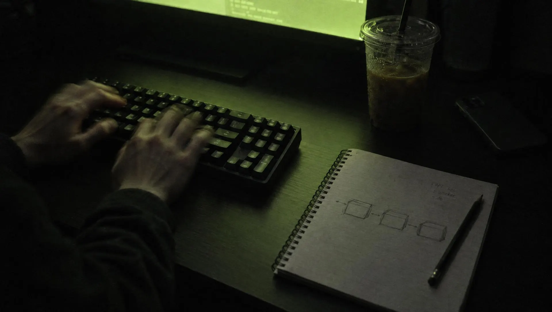 A developer's hands at a keyboard at night, lit by a green terminal glow, a notebook of sketches beside