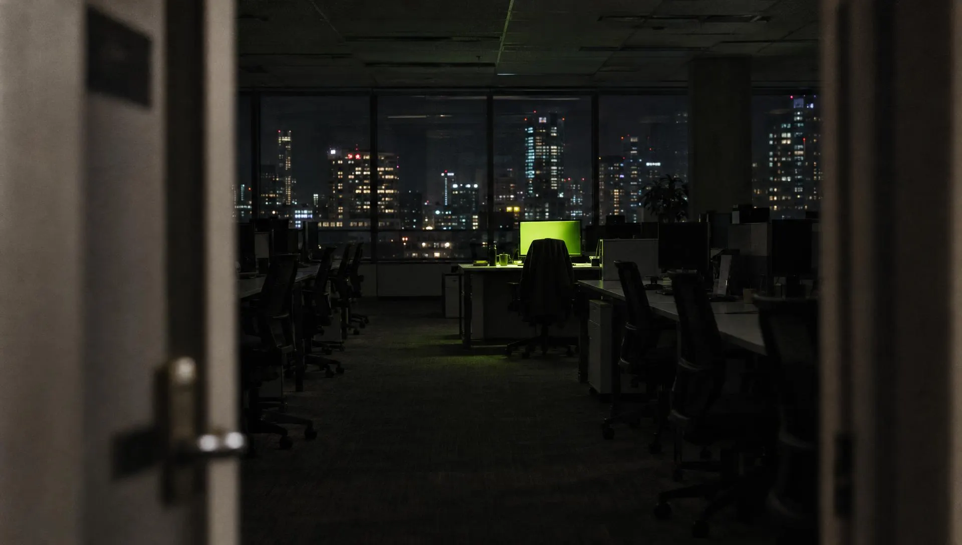 An empty office floor at night, one desk still lit green, chairs pushed in