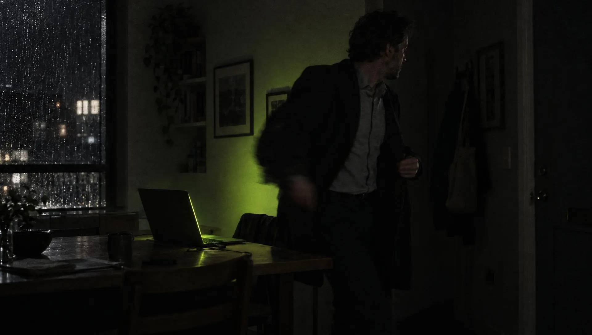 A man at a kitchen table at night stepping away from a laptop that keeps working, green screen glow on the wall