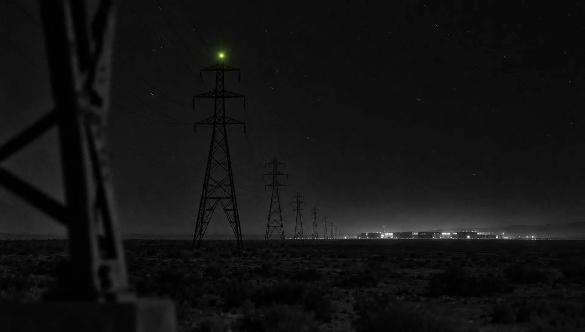 High-voltage pylons crossing a desert at night toward a lit data-centre campus, one green warning light