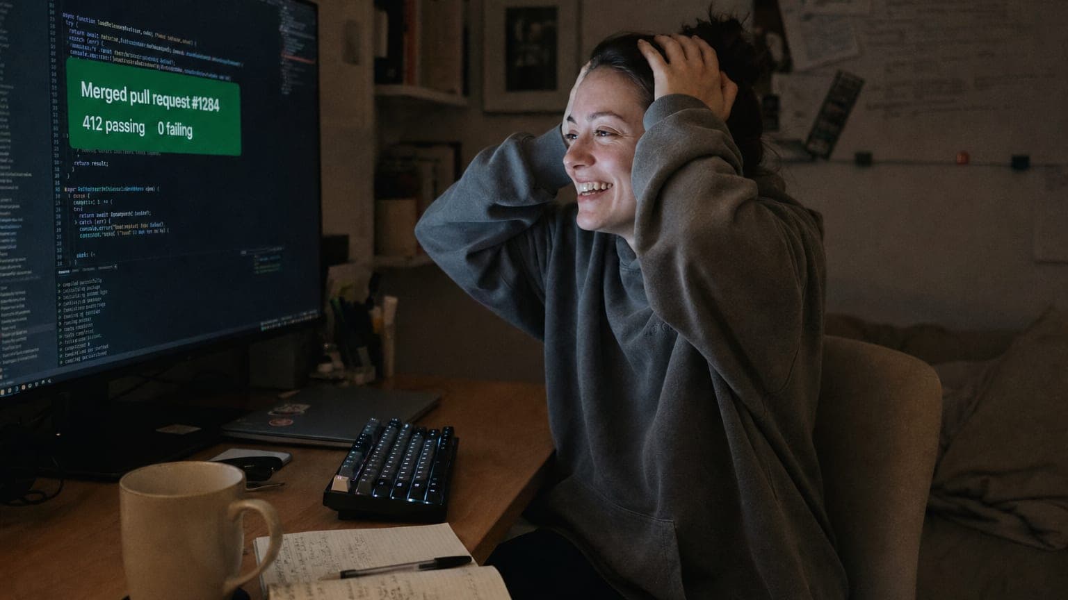 A developer leaning back from the keyboard, hands behind head, laughing in relief as a screen shows a merged pull request and passing tests.