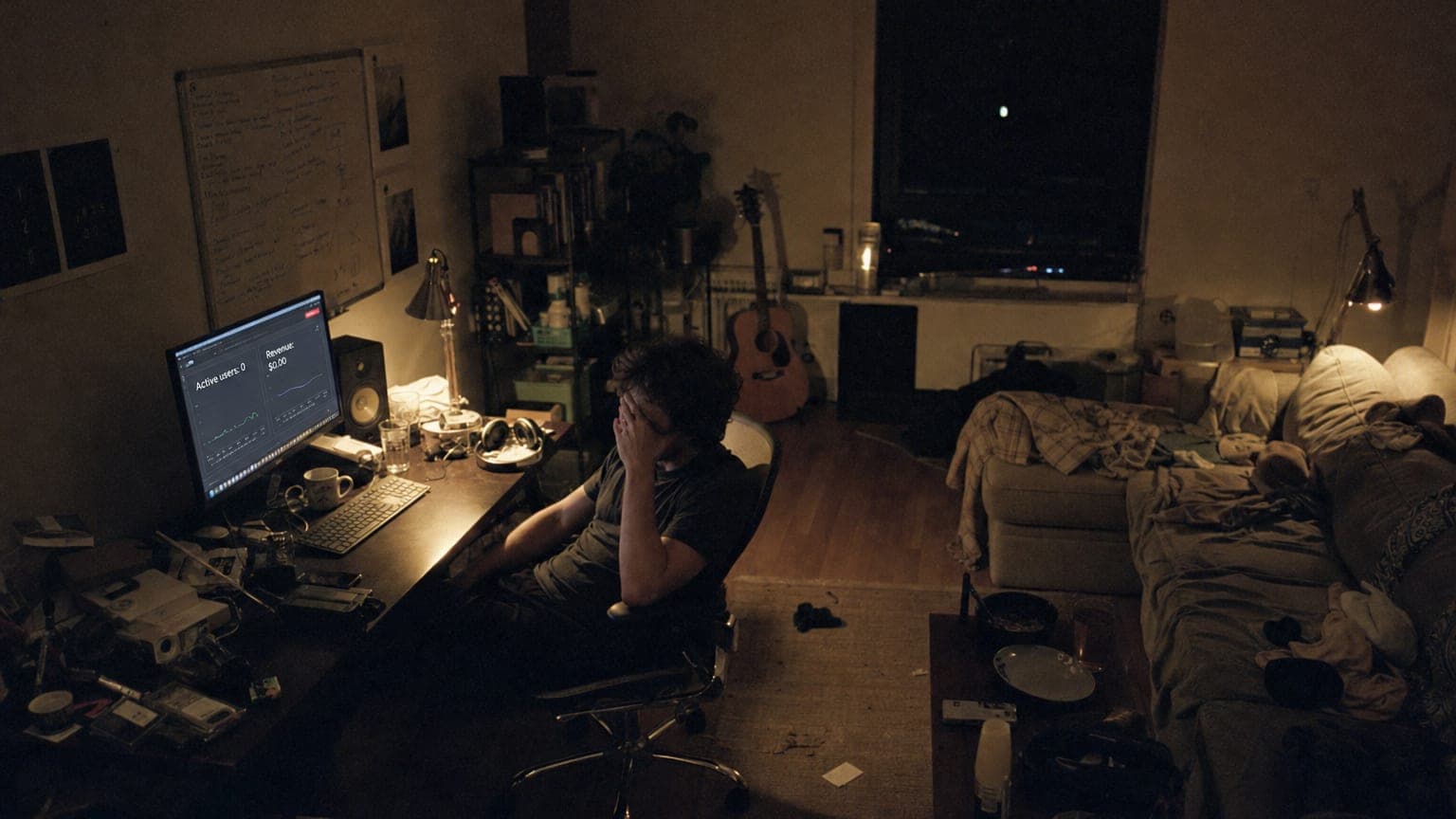 A wide, dark, cluttered solo studio in the dead hours, a builder slumped at the desk, monitors ignored.