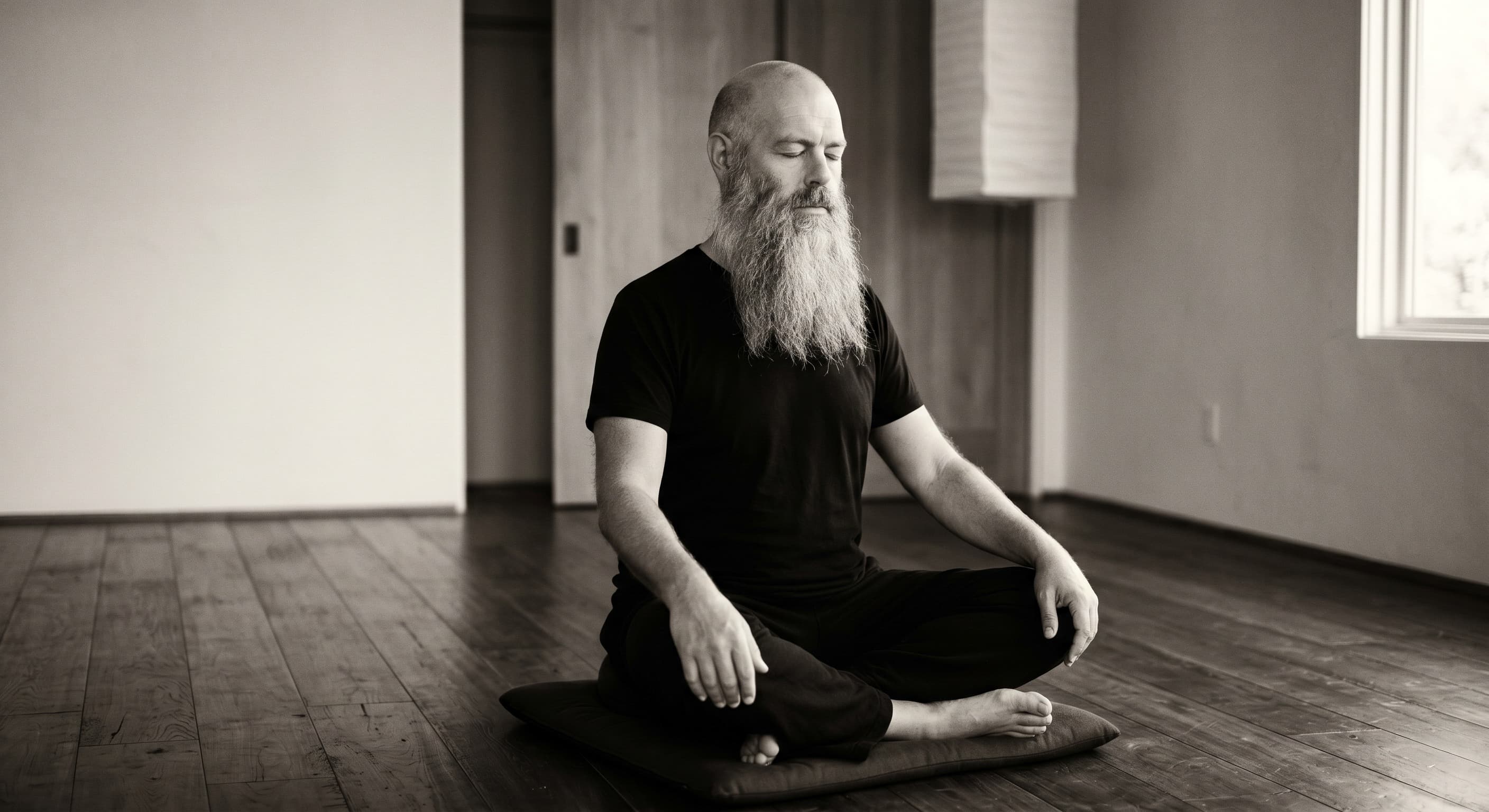 A bearded figure meditating on a zabuton in an empty wooden room.